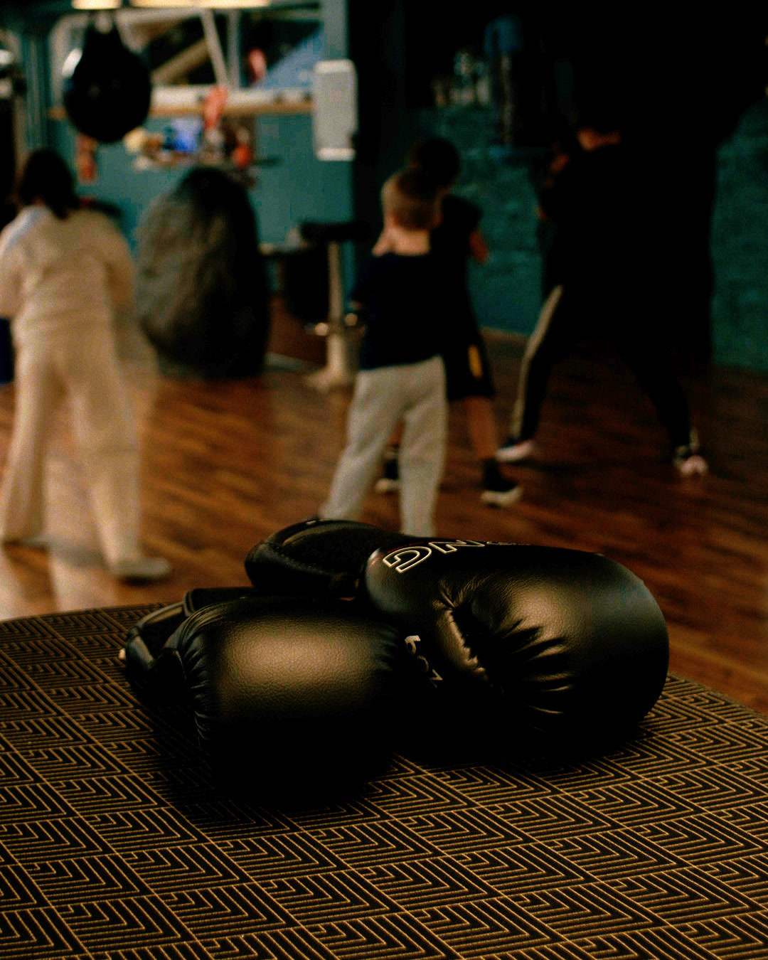 Boxing gloves lie on a mat, with kids training in the background.