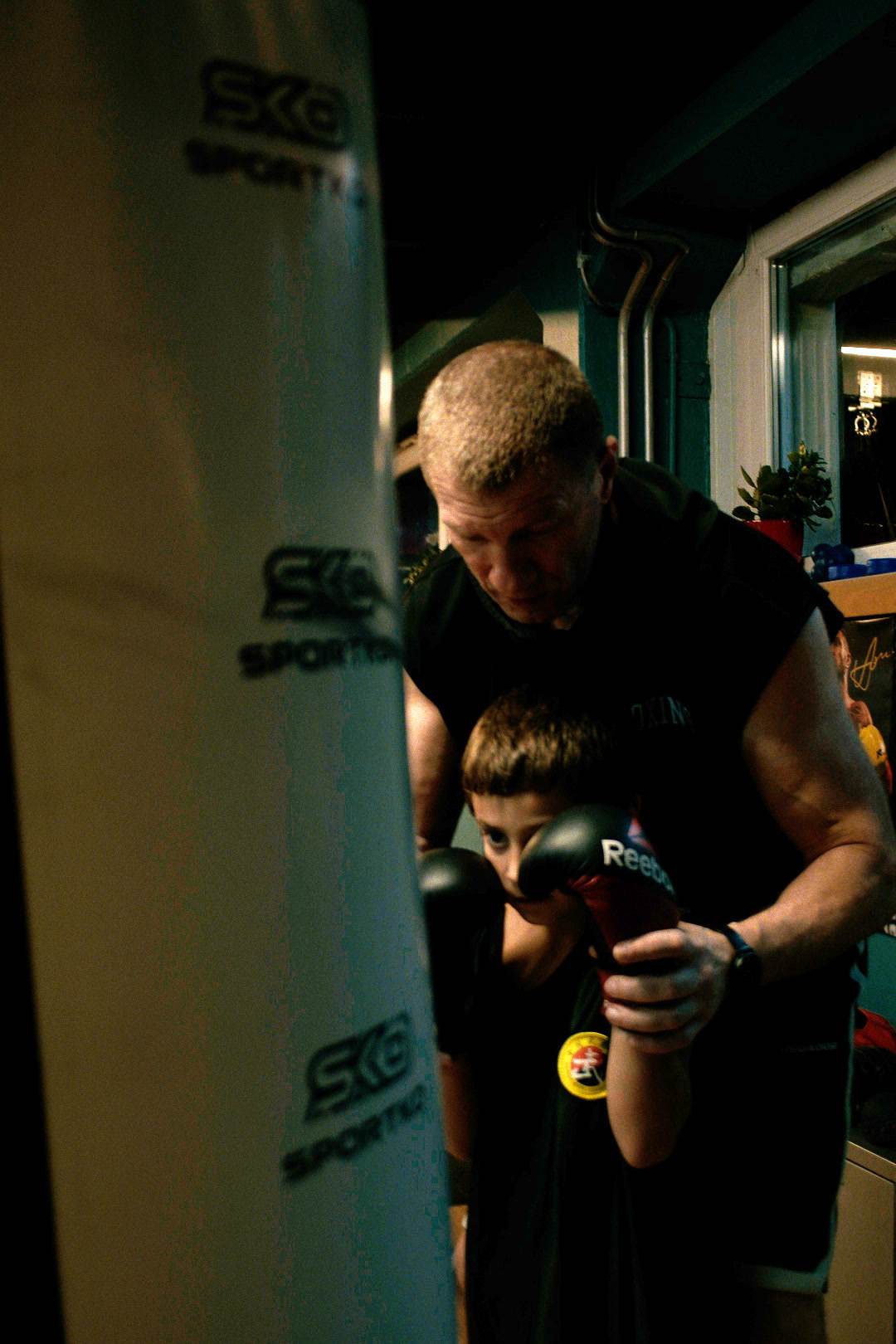 The coach leans down to a boy at the heavy bag and shows him how to hold his fists.